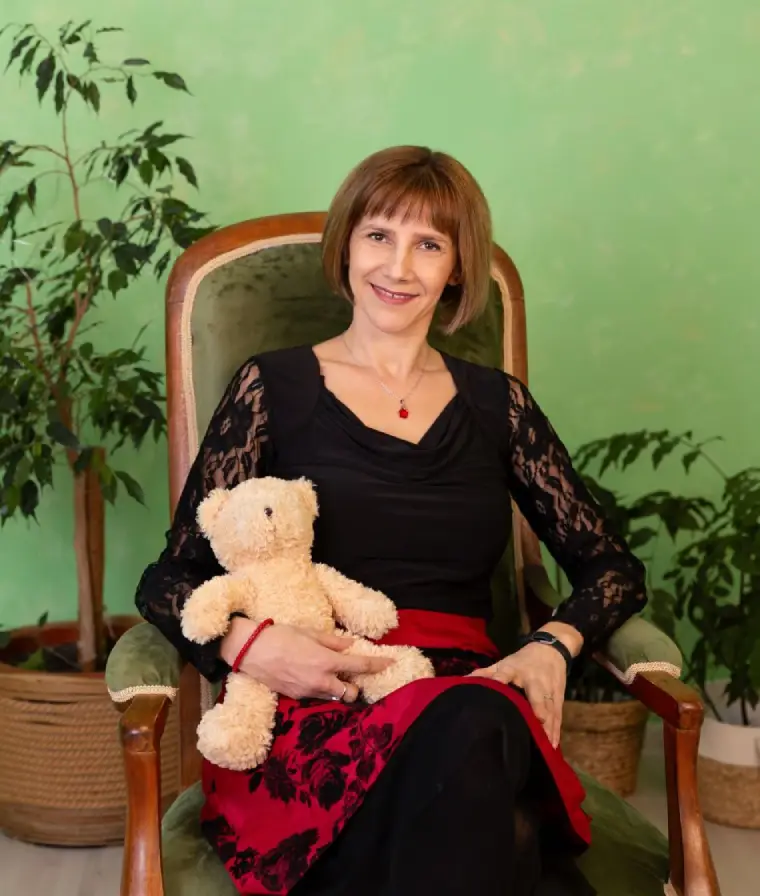 Carmen Liebert, baby sleep coach, sitting in a vintage armchair holding a teddy bear
